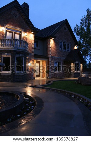 view of a large luxurious home in the evening after a light rain with the house lit up