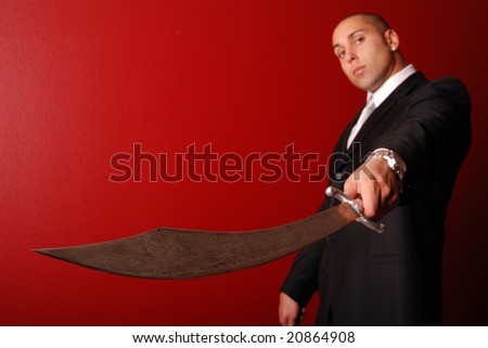 Man in business suit against a red background holding a samurai sword out towards the camera. Focus is on the detailed pattern on the end of the sword.
