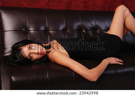 Beautiful Hispanic woman wearing a black dress laying on her back on a brown leather sofa.