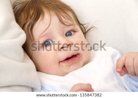 Picture shows a baby. Baby is eating and smiling. He is looking to camera. White background.