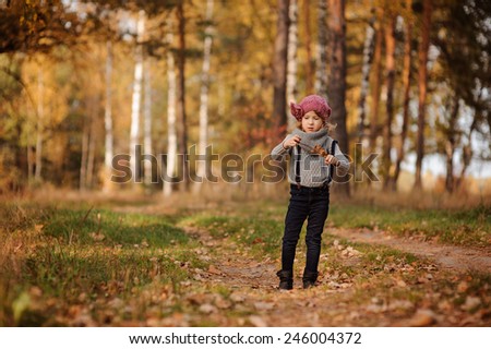 adorable child girl in pink hat and grey sweater plays with stick and leaves on the walk in autumn forest