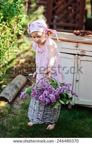 child girl in pink plaid dress standing near white vintage bureau and holding basket of lilacs in spring garden