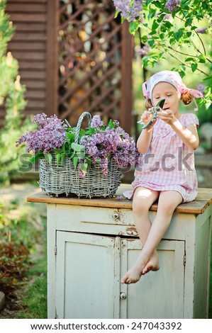 spring portrait of child girl in pink plaid dress cutting lilacs with secateurs in sunny garden