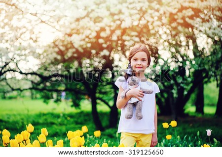 adorable smiling child girl with teddy bear on the walk in spring park with blooming tulips and apple tree on background