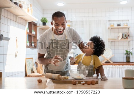 Cheerful smiling Black son enjoying playing with his father while doing bakery at home. Playful African family having fun cooking baking cake or cookies in kitchen together. Single Dad Lifestyle