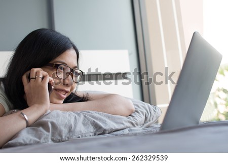 Asian girl is talking with a other person with her cell phone and laptop on a bed in her vacation at her house.