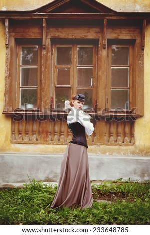 Beautiful redhair woman in vintage clothes standing near old ancient house with wooden window. Toned in warm vintage colors.