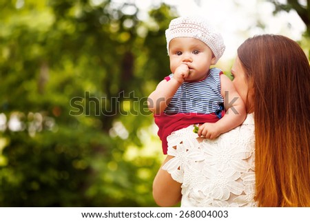Cute Toddler Baby Girl sitting on Mom`s Hands, looking into camera. Relaxing in green summer Park.