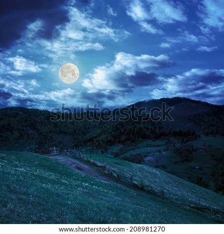 summer landscape. fence near the meadow path to village on the hillside. forest with lumber on the mountain at night in full moon light