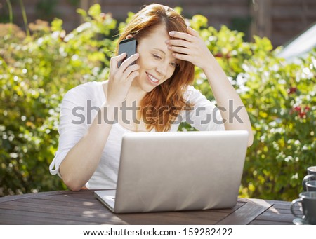 Smiling young redhead woman sitting at a wooden table in the garden in front of her laptop computer chatting on her mobile phone in the summer sunshine in front of green leaves