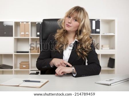 Manageress sitting at her desk in the office pointing to her watch making a point that someone is late for an interview of meeting with her