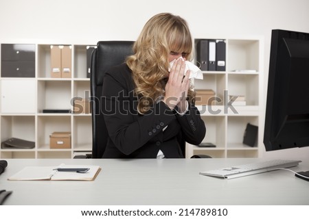 Businesswoman suffering from a cold or hay fever blowing her nose at her desk with a tissue as she continues reading the screen of her computer