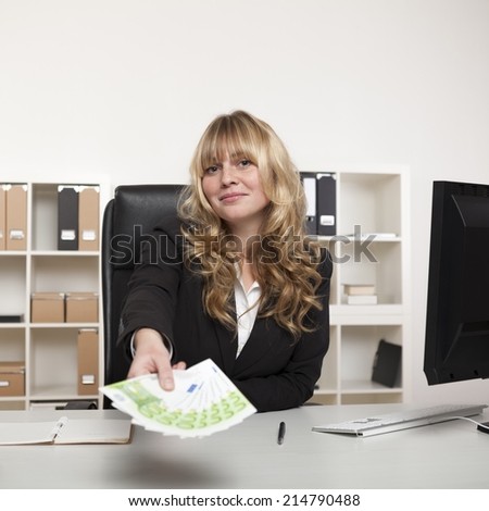 Beautiful young businesswoman holding out a fistful of banknotes as she stretches across her desk conceptual of success, a bribe or pay-off