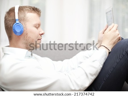 Man immersed in his music listening with closed eyes as he relaxes on a sofa with his feet up holding a tablet or MP3 storage device attached to his headphones