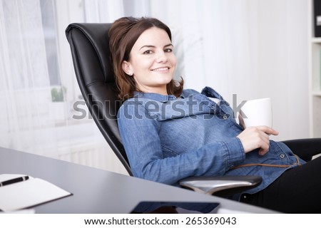 Relaxed businesswoman enjoying her coffee break reclining back in her chair at the desk looking at the camera with a lovely friendly warm smile