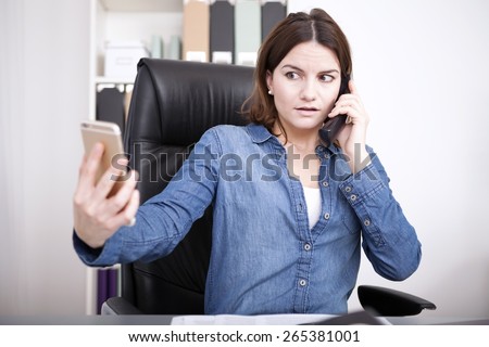 Businesswoman talking on the land line telephone phone in the office while simultaneously checking her mobile with a worried expression