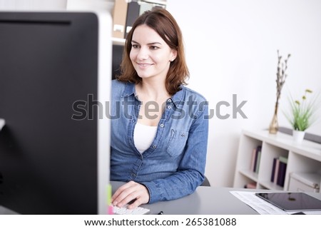 Businesswoman smiling as she works at her desktop while sitting at her desk, view past the edge of the monitor