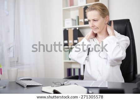 Tired Female Medical Doctor Sitting at her Office and Massaging the Back of her Neck While Looking Down.