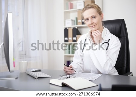 Smiling Blond Female Doctor Sitting at her Table, with Pen and Paper to Make Diagnosis, and Looking at the Camera with Hand on her Chin.