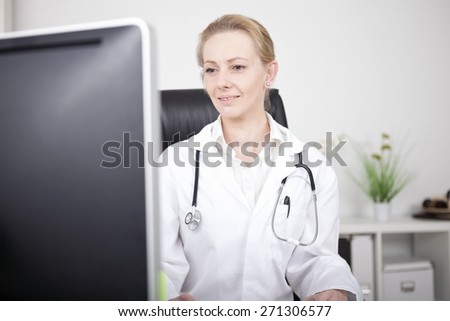 Close up Blond Woman Doctor with Stethoscope Apparatus on her Shoulders Sitting at her Office While Facing the Computer Monitor.