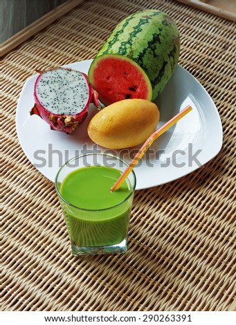 Blended green smoothie and sliced watermelon, mango and dragon fruit on white plate on rattan table, view 1