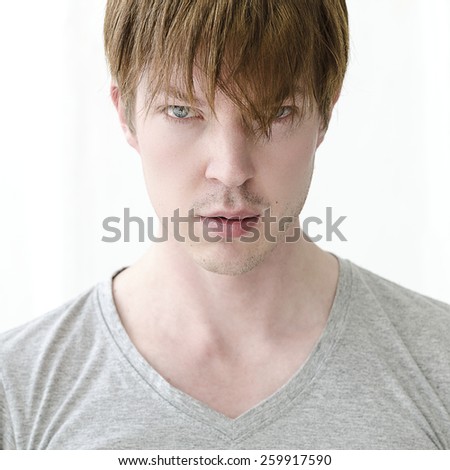 A square format photographic headshot image of a young blue eyed man wearing a grey t-shirt photographed against a white background