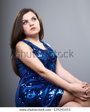 Attractive young woman in a blue shiny dress. Woman sitting and looking in the upper-left corner. On a gray background