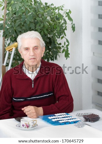 Senior man with walking stick sitting at table with pills container and cup of coffee on it