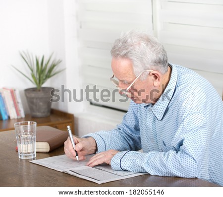 Senior man in pajamas with reading glasses sitting at table and solving crosswords