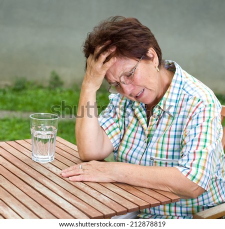 Senior woman suffers from headache, sitting at table with glass of water in front of her