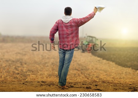 Excited young farmer waving with bank loan forms in hands in front of working tractor in field