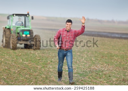 Happy young farmer waving with hand on farmland with tractor in background