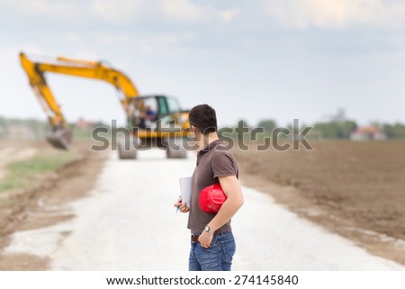 Engineer holding helmet and looking behind at excavator on road construction site