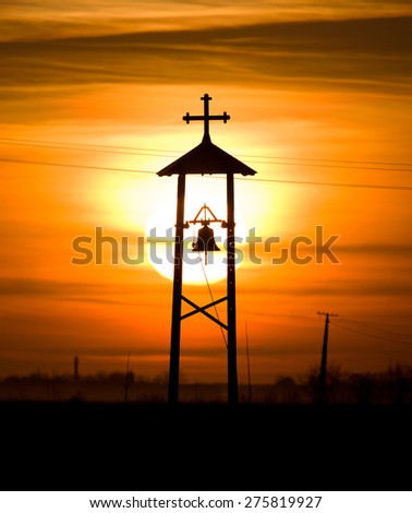 Dusk scene of Christian bell and cross silhouette on meadow with sun in background