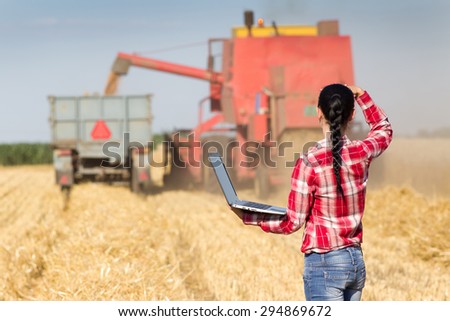 Young beautiful woman in plaid shirt standing with laptop in wheat field and looking at combine harvester and tractor trailer
