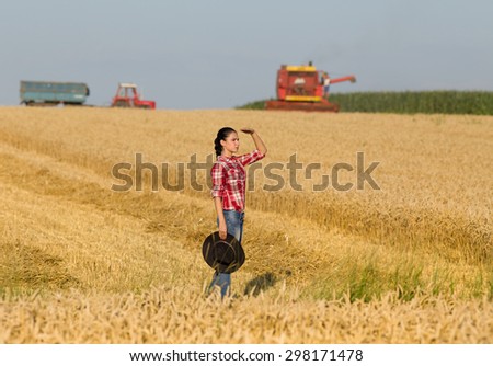 Beautiful young girl with cowboy hat  looking far away on wheat field, combine harvester and tractor with trailer working in background