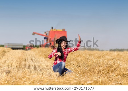 Beautiful young girl with cowboy hat squatting in harvested whet field, combine harvester and tractor with trailer working in background