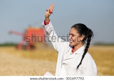 Young happy woman agronomist standing on wheat field and waving hand. Combine harvester working in background