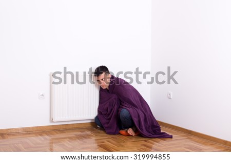 Young man covered with blanket sitting on floor beside radiator and listening if it works