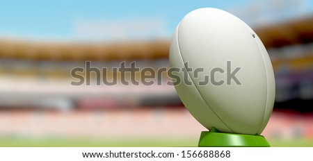 A plain white textured rugby ball on a green kicking tee in a stadium
