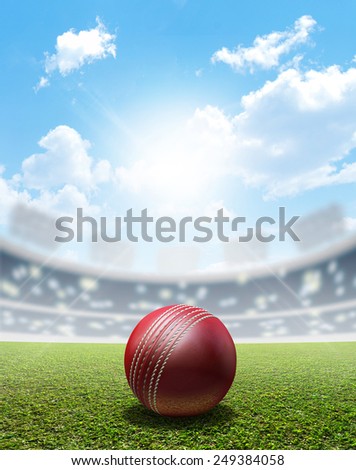 A cricket stadium with a red leather cricket ball on an unmarked green grass pitch in the daytime under a blue sky