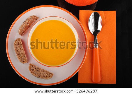 bowl of pumpkin hokkaido soup with bread  on white plate on black acrylic  sheet, orange spoon on orange paper napkin and pumpkin hokkaido