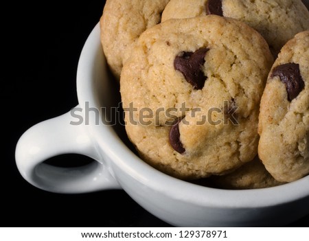 A close-up photograph of soft, homemade chocolate chip cookies in a white coffee cup on a black background.