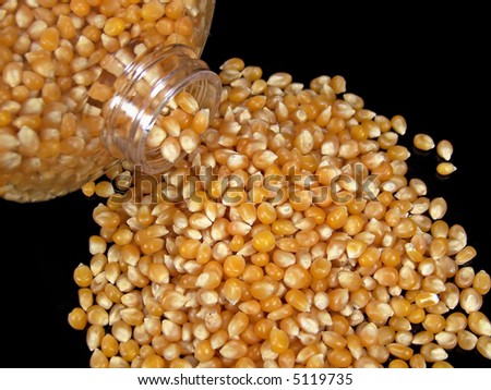 Color photograph of popcorn kernels pouring onto a black table from a clear plastic container.