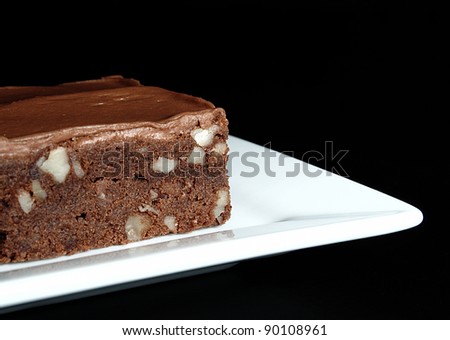 A moist, fudge chocolate brownie with nuts and chocolate icing on a square white plate on a black background.