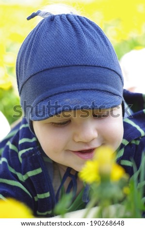 Beautiful happy little baby boy in hat sitting on a green meadow with yellow flowers dandelions on the nature in the park