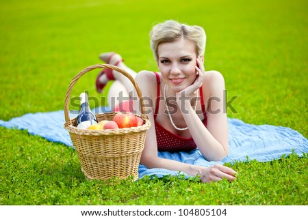 Young blond woman is lying on grass. Basket with bread, fruit and bottle of vine is standing near her