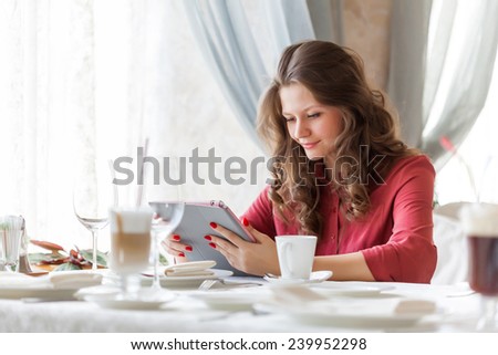 Young smiling woman is drinking coffee in a cafe and holding a tablet computer in hands