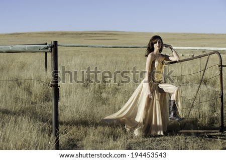 Sexy brunette woman wearing a gold evening dress with gum boots, posing outside in a field in the sun against a rusty metal farm gate.