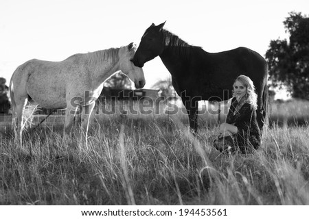 Black and white portrait of blonde woman seated next to two horses in a field